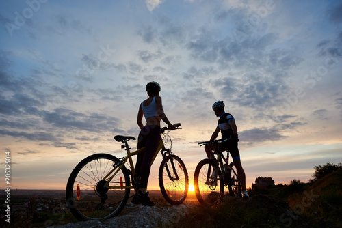 Two young sports people on mountain bikes stand on rock on top of hill looking at the sun at sunset. Girl and guy dressed in helmets and sportswear stand facing each other under evening sky