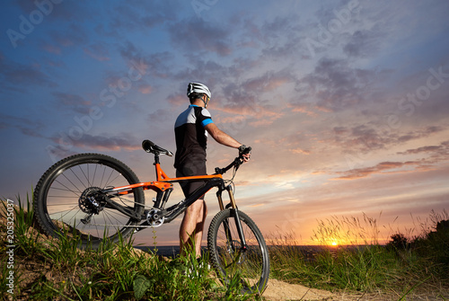 Back view of cyclist male in a helmet and sports clothes rests on the background of a sunset from a height watching the mountain in the distance, the enchanting sky and the bright sun