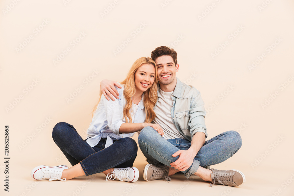 Image of nice and cute couple in denim clothing smiling and hugging together while sitting on the floor with legs crossed, isolated over beige background Image of nice and cute couple in denim clothing smiling and hugging together while sitting on the floor with legs crossed, isolated over beige background