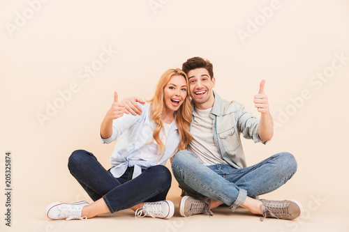 Image of handsome man and beautiful woman hugging together while sitting on the floor with legs crossed and showing thumbs up, isolated over beige background
