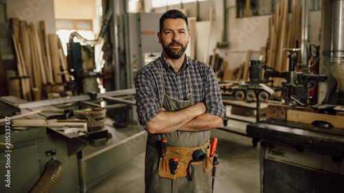 Portrait of a smiling young woodworker standing with his arms crossed by a bench saw in his workshop full of carpentry equipment