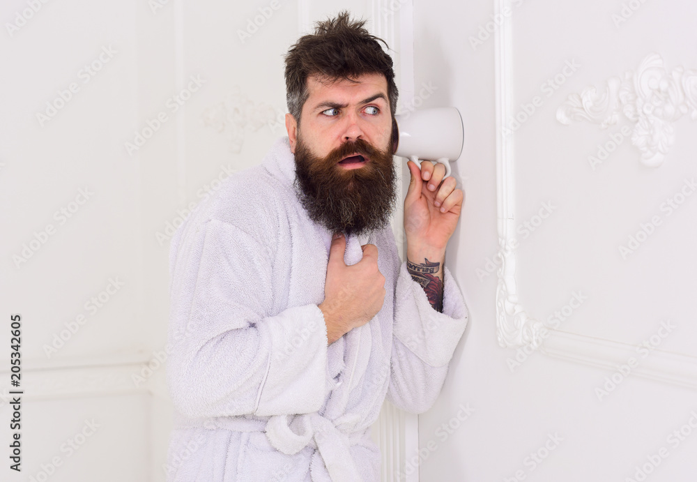 Man in white interior spying, eavesdropping. Secret and spy concept. Man with beard and mustache eavesdrops using mug near wall. Hipster in bathrobe on strict face secretly listen conversation.