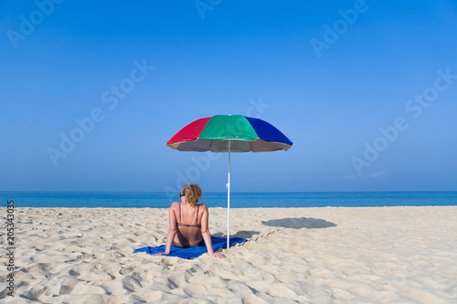 A lonely woman is sitting on the beach next to a beach umbrella.