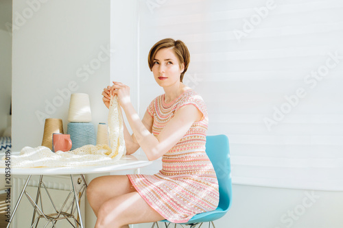 Young short-haired woman knitting dress for her hobby sitting in kitchen with coffee cup in sunny morning. Business crochet handmade home concept.