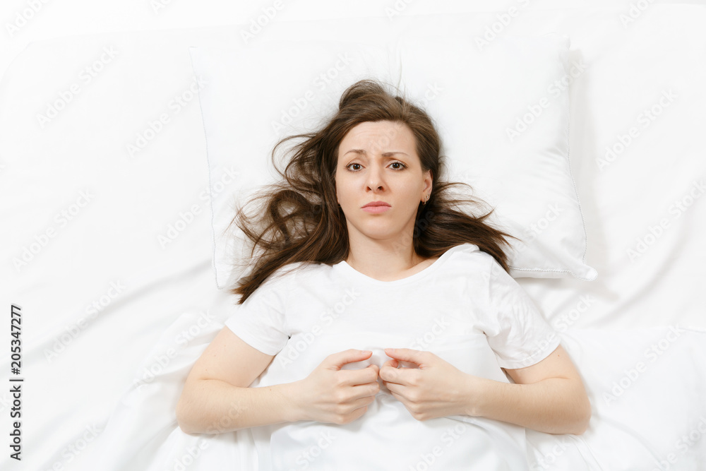 Top view of tired stressed crying young woman lying in bed with white sheet, pillow, blanket. Pensive frustrated sad upset female spending time in room. Rest, relax, bad mood concept. Copy space. Top view of tired stressed crying young woman lying in bed with white sheet, pillow, blanket. Pensive frustrated sad upset female spending time in room. Rest, relax, bad mood concept. Copy space.