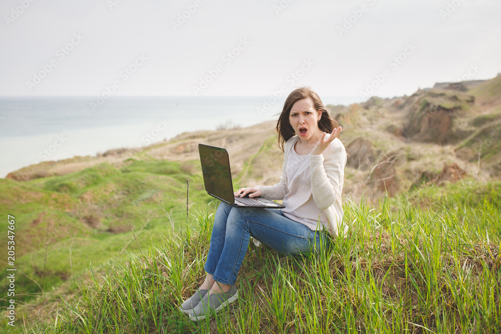 Young irritated dissatisfied business woman or student in casual clothes sitting on grass using laptop in field spreading hands working outdoors on green background. Mobile Office. Lifestyle concept.