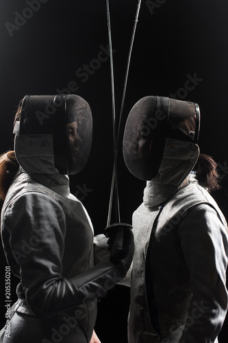 two women wearing helmets and white uniforms fencing on black background