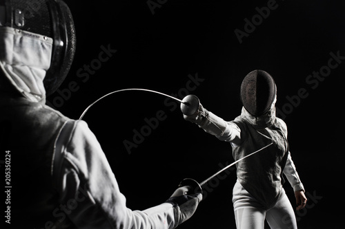 Slika na platnu two women wearing helmets and white uniforms fencing on black background