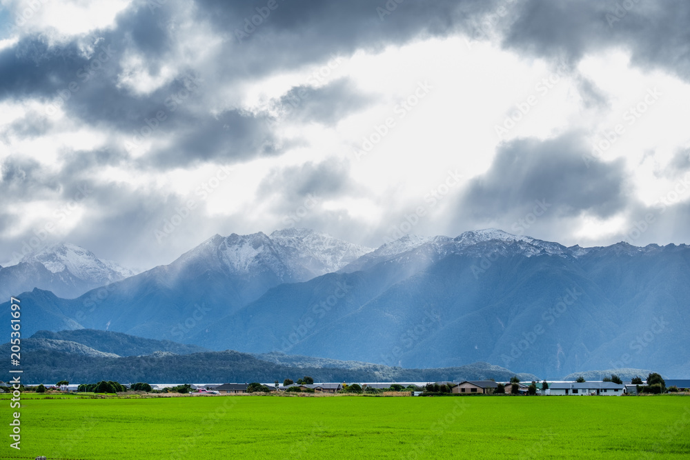 A stunning landscape scene of a rural area with green grassland cloudy snow mountain and sun rays after raining day.