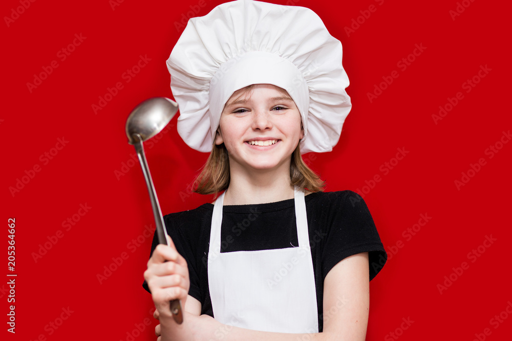 Happy little girl in chef uniform holds spoon isolated on red. Kid chef. Cooking Process Concept Happy little girl in chef uniform holds spoon isolated on red. Kid chef. Cooking Process Concept