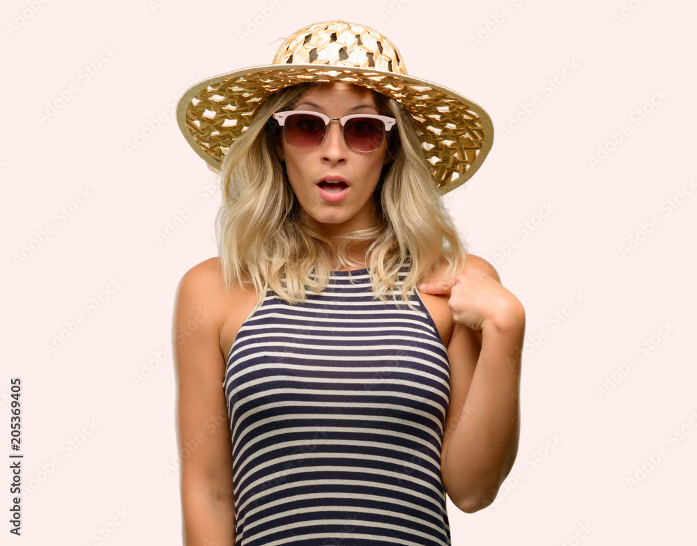 Young woman using sunglasses wearing summer hat happy and surprised cheering expressing wow gesture, pointing with finger Young woman using sunglasses wearing summer hat happy and surprised cheering expressing wow gesture, pointing with finger