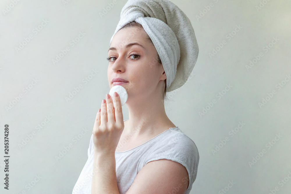 Beauty portrait of a smiling brunette woman with towel on head with soft healthy skin removing make up with cotton pad isolated over white background. Skincare cleansing spa relax concept Beauty portrait of a smiling brunette woman with towel on head with soft healthy skin removing make up with cotton pad isolated over white background. Skincare cleansing spa relax concept