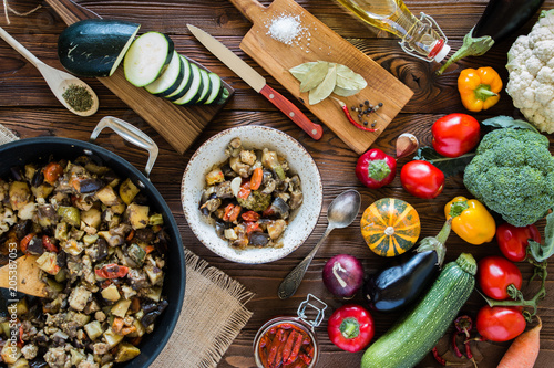 vegetable stew in the pan and plate and fresh vegetables ingredients on wooden table, top view