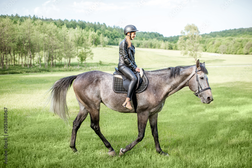 Woman in leather jacket with protective helmet riding a horse on the green meadow Woman in leather jacket with protective helmet riding a horse on the green meadow