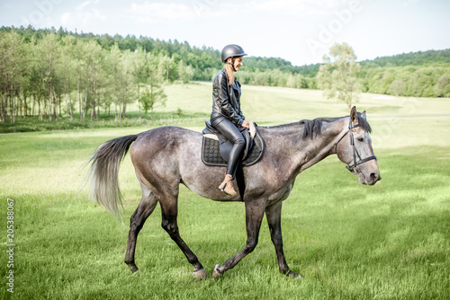 Woman in leather jacket with protective helmet riding a horse on the green meadow © rh2010