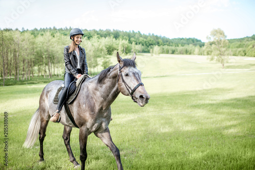 Woman in leather jacket with protective helmet riding a horse on the green meadow