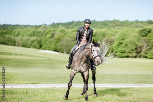 Handsome man in leather jacket with protective helmet riding a horse on the green meadow