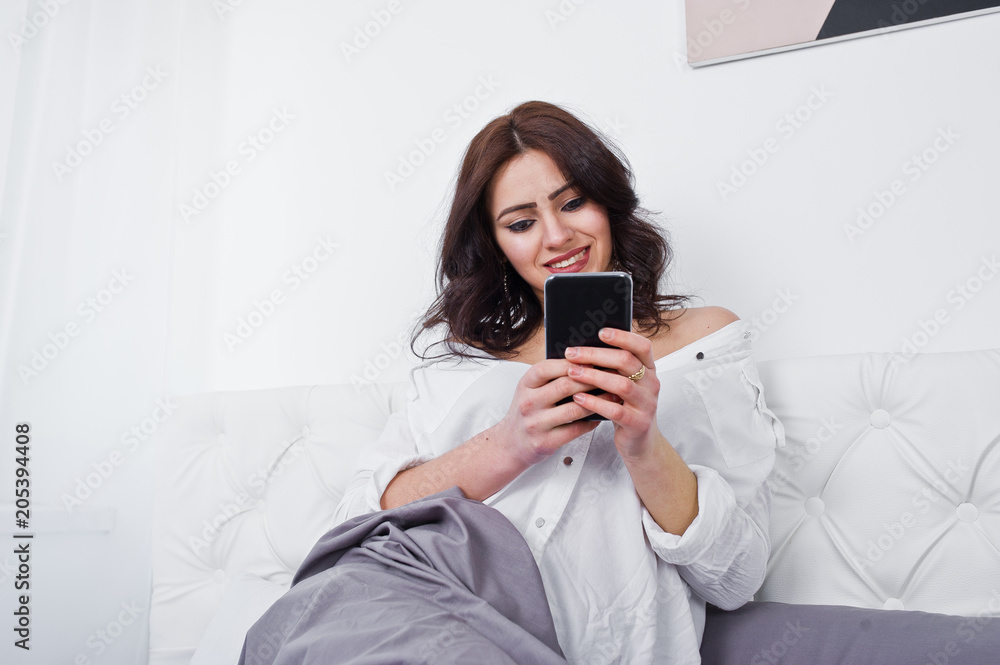 Studio portrait of brunette girl in white blouse lying on bed and using mobile phone. Studio portrait of brunette girl in white blouse lying on bed and using mobile phone.