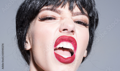 Woman with attractive red lips shouting. Lady in black wig with make up on grey background. Scandalous lady concept. Girl on scandalous shouting face, close up. photo