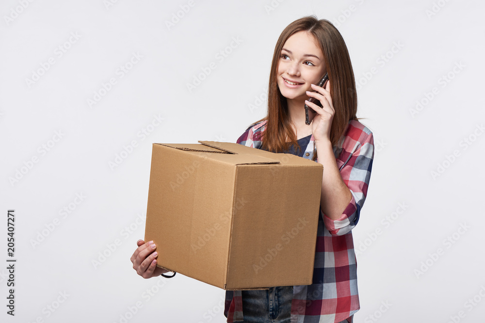 Delivery, relocation and unpacking. Smiling young woman holding cardboard box and talking on cellphone looking away at blank copy space. Call center and customer support service concept. Delivery, relocation and unpacking. Smiling young woman holding cardboard box and talking on cellphone looking away at blank copy space. Call center and customer support service concept.