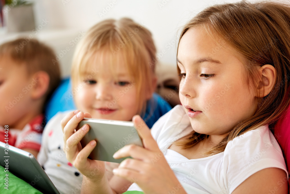childhood, technology and family concept - happy little kids with smartphone and tablet pc computer in bed at home childhood, technology and family concept - happy little kids with smartphone and tablet pc computer in bed at home