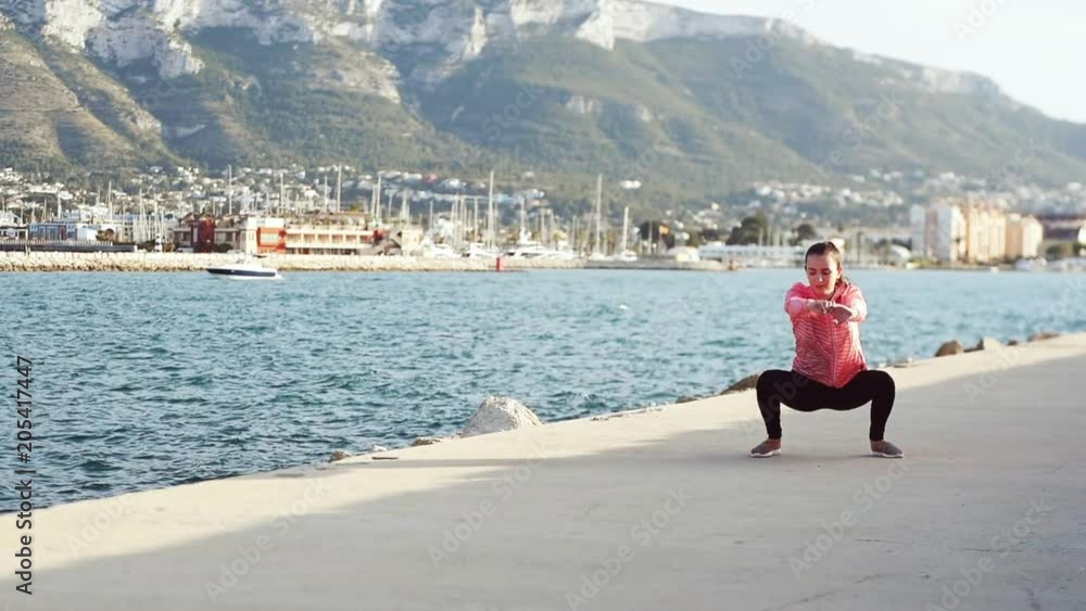 Young sporty woman doing jump squats by the sea with mountain on background on a sunny day. Young sporty woman doing jump squats by the sea with mountain on background on a sunny day.