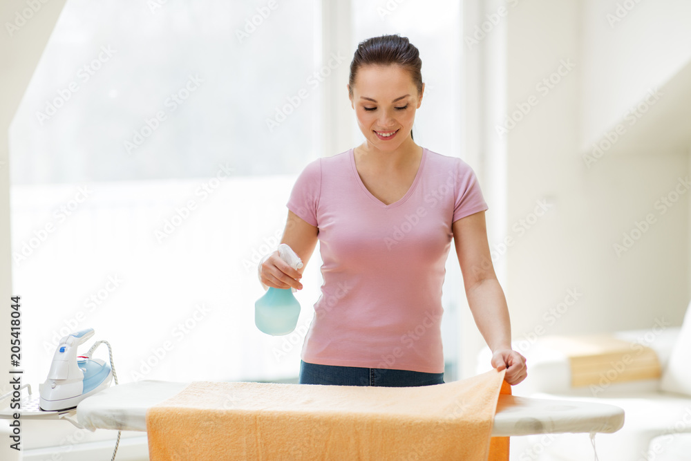 housework and household concept - happy woman or housewife spraying bath towel on ironing board at home housework and household concept - happy woman or housewife spraying bath towel on ironing board at home