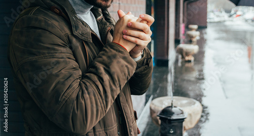 handsome man standing on the street in winter time and warming his hands. cold day. it's raining. man in jacket. Town architecture on the background. Outdoors