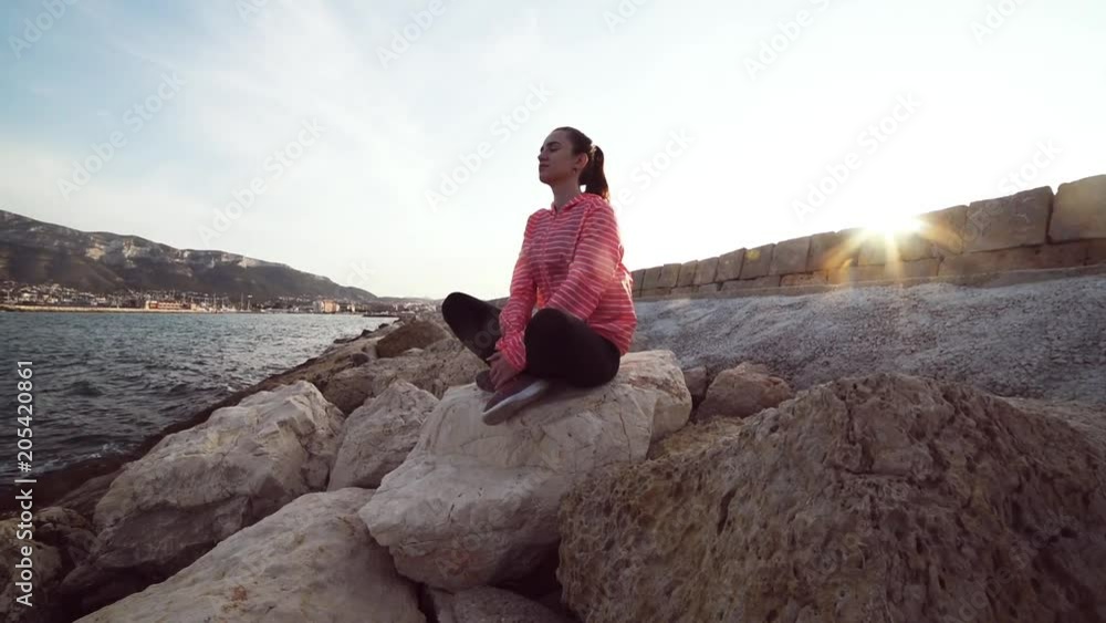 Young woman sitting in lotus position slow motion at sunset sea. Girl practicing meditation on the beach. Waves rolling into rocking shore. Young woman sitting in lotus position slow motion at sunset sea. Girl practicing meditation on the beach. Waves rolling into rocking shore.