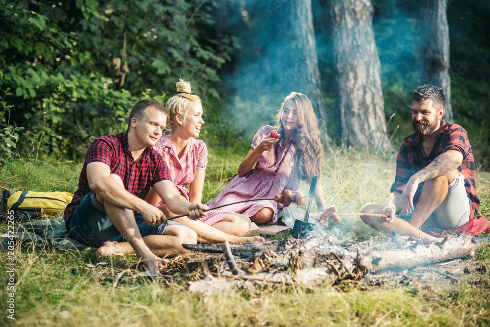Group of friends camping in woods. Two couples cooking sausages over fire. Brunette and blond girls relaxing with their boyfriends outdoors