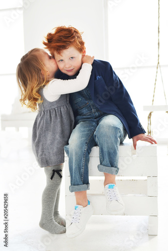 Sister kissing her brother in the cheek. Cute blond baby girl and red boy brother in the white studio. Love in the family photo