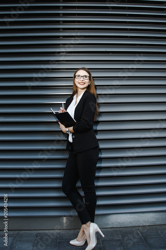 A young woman in a business suit is near the office. Business woman in glasses and a notebook. The girl is making notes on her daily party. Good girl smiles. The lady stands near a gray wall.