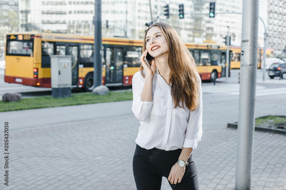 A business woman speaks through the phone. A good girl is sitting on the bench. A young woman in the city center.