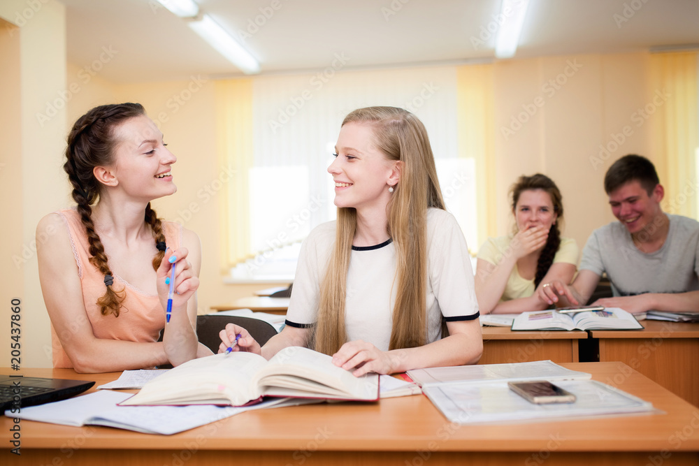 Two happy students learning on line together with a computer in a classroom with other classmate in the background Two happy students learning on line together with a computer in a classroom with other classmate in the background
