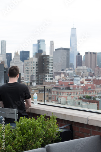 Man sits at the bar overlooking Lower Manhattan drinking a bottled water on a trendy rooftop bar in New York. Man in black shirt looking out at New York City from a rooftop bar.