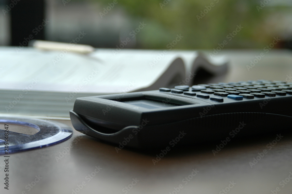 Math still life photos. Calculator, protractor, open book, and wooden pencil on a brown desk next to a window in a library at university. Studying math and doing homework at school. Math still life photos. Calculator, protractor, open book, and wooden pencil on a brown desk next to a window in a library at university. Studying math and doing homework at school.