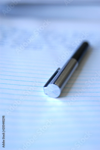 Still life of pen and paper. Macro lens, shallow depth of field focus on the tip of the pen. Silver ball point pen laying on a piece of white college ruled paper with blue lines, writing on the page.