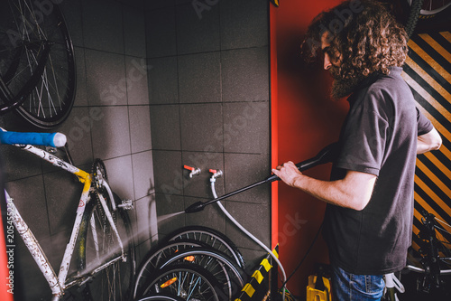 Services professional washing of a bicycle in the workshop. Close-up of hand Young Caucasian stylish man doing bicycle cleaning using automatic electric water pump. Sprays scatter from the pressure