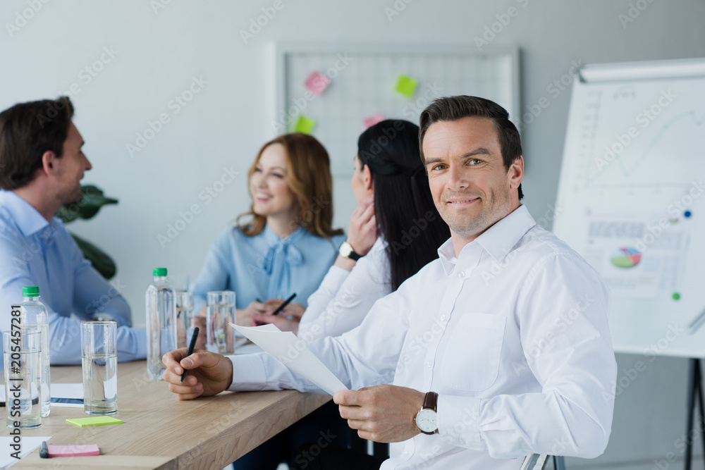 Fototapeta premium selective focus of smiling businessman with papers and colleagues at workplace in office