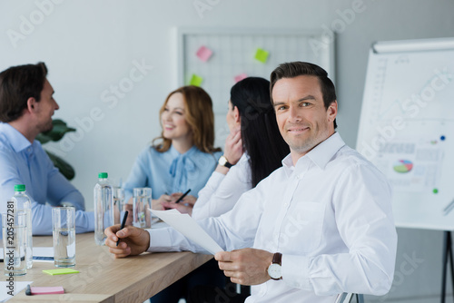 selective focus of smiling businessman with papers and colleagues at workplace in office