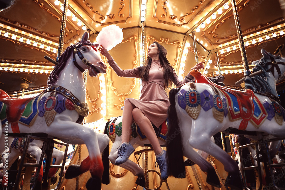 Young beauty model woman posing with old horse carousel in summer park with magic lights