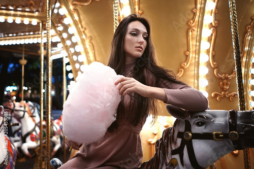 Young beauty model woman posing with old horse carousel in summer park with magic lights