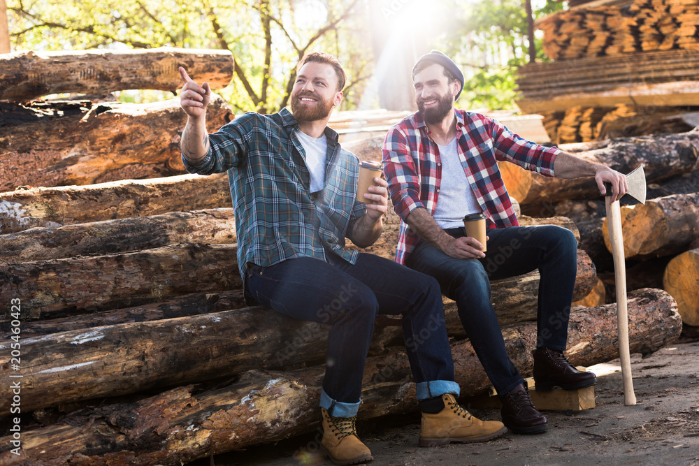 smiling lumberjack with coffee cup pointing by finger to partner with axe sitting on logs at sawmill