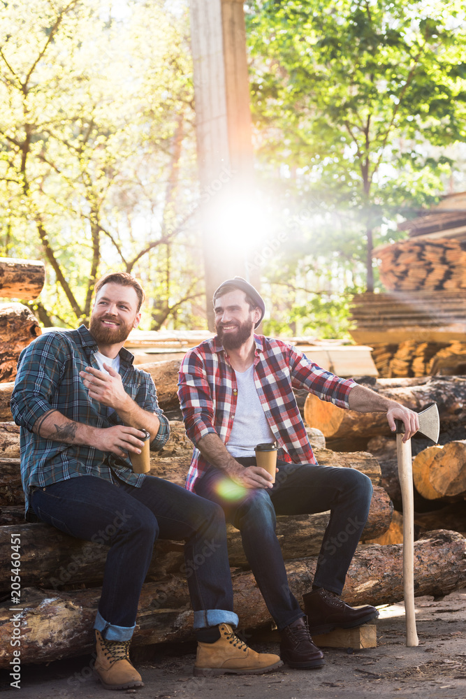 lumberjack with coffee cup pointing by finger to partner with axe sitting on logs at sawmill lumberjack with coffee cup pointing by finger to partner with axe sitting on logs at sawmill