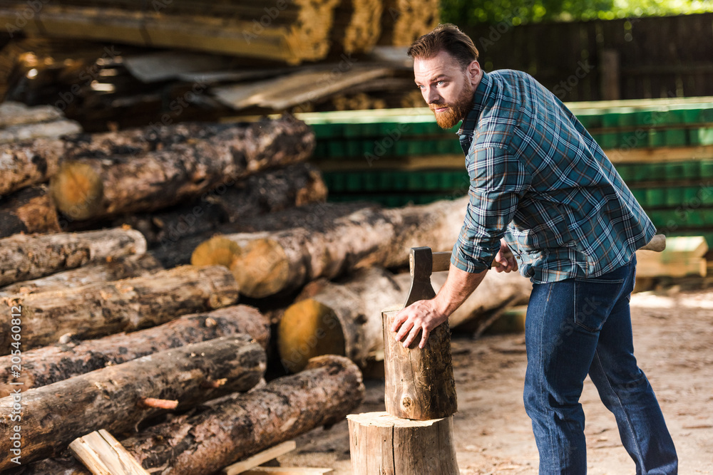 bearded lumberjack in checkered shirt chopping log at sawmill