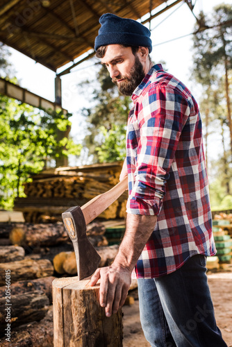 bearded lumberjack in checkered shirt preparing to chop log at sawmill