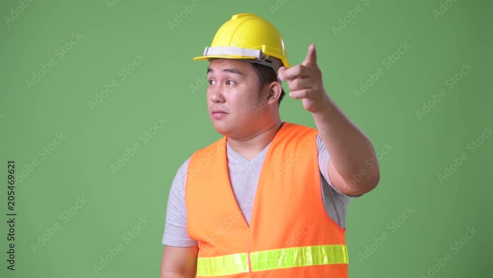 Young handsome overweight Asian man construction worker against green background Young handsome overweight Asian man construction worker against green background