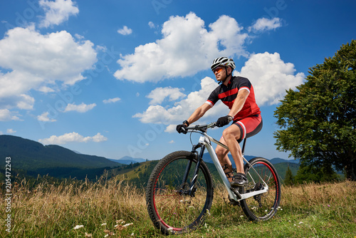 Professional sportsman rider in sportswear and helmet cycling bicycle in high grass, male cyclist enjoying view of Carpathian mountains on sunny summer day. Active lifestyle and extreme sport concept