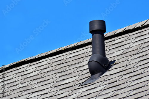 Black air ventilation chimney on grey shingles roof of residental house on blue sky background with copy space for text.