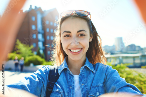 young smiling attractive woman in jeans clothes makes selfie on smartphone at sunny day on the cityscape background. woman posing in city scape.